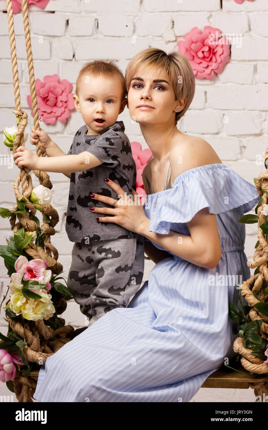 Attractive young mother with her small son on swing on light brick`s background Stock Photo